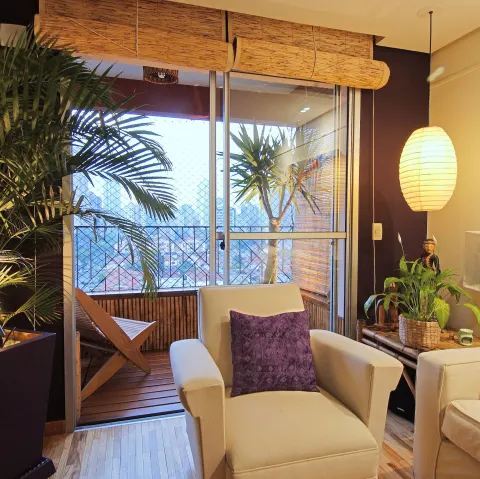 Sala com poltrona clara, almofada roxa e varanda com plantas